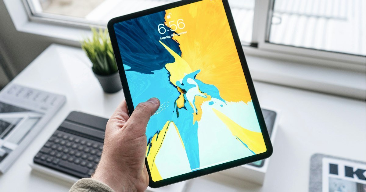 A hand holding an iPad Pro showing its colorful lock screen, with a keyboard and plant on the desk behind